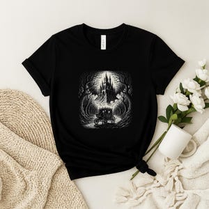 May include: Black t-shirt with a monochrome illustration of a horse-drawn carriage on a road leading to a castle. The design includes trees and a detailed castle silhouette, creating a gothic or fantasy theme.