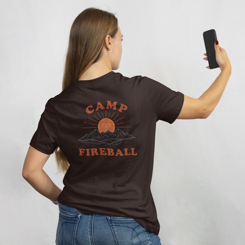 K&ouml;nnte beinhalten: Dunkelbraunes T-Shirt mit den Worten "CAMP FIREBALL" in orangefarbener Schrift. Das Design zeigt eine Sonne, Berge und eine geometrische Form. Das T-Shirt wird von einer Person gehalten.