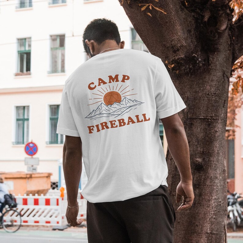 K&ouml;nnte beinhalten: Wei&szlig;es T-Shirt mit einem orangefarbenen Aufdruck auf dem R&uuml;cken. Das Design enth&auml;lt die Worte "CAMP FIREBALL", eine Sonne, Berge und eine geometrische Form. Das T-Shirt ist kurz&auml;rmelig.