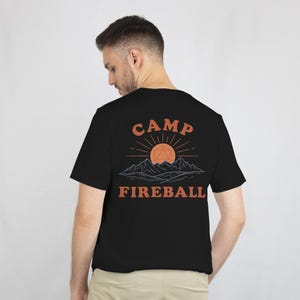 K&ouml;nnte beinhalten: Schwarzes T-Shirt mit einem grafischen Design auf der R&uuml;ckseite. Das Design zeigt die Worte "CAMP FIREBALL" in Orange, mit einer Sonne und einer Bergillustration &uuml;ber dem Text. Das Shirt ist aus weichem Material.