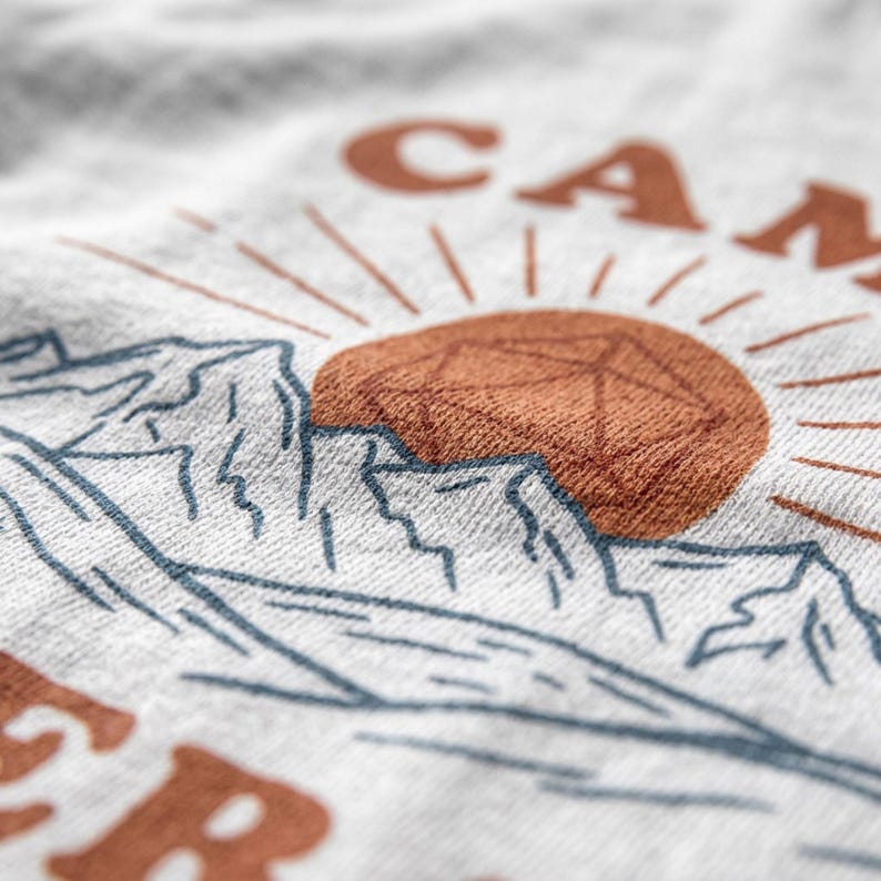 K&ouml;nnte beinhalten: Nahaufnahme eines hellgrauen T-Shirts mit einem Berg- und Sonnengrafik. Das Design zeigt eine braune Sonne mit Strahlen, blaue Berggipfel und das Wort "CAMP" in Braun. Das T-Shirt hat ein weiches, strukturiertes Aussehen.