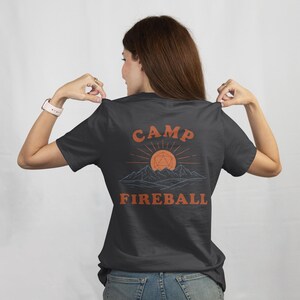 K&ouml;nnte beinhalten: Dunkelgraues T-Shirt mit den Worten "CAMP FIREBALL" in Orange, mit einer Berg- und Sonnengrafik. Das Design enth&auml;lt eine geometrische Form in der Mitte der Sonne. Ein l&auml;ssiges T-Shirt f&uuml;r Camping- und Gaming-Enthusiasten.