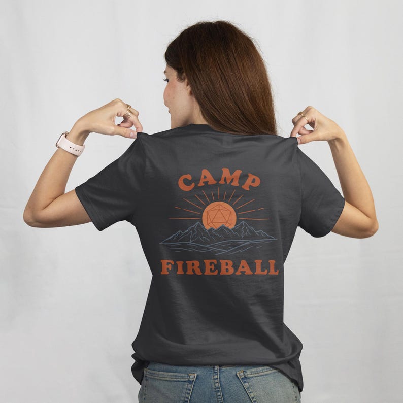 K&ouml;nnte beinhalten: Dunkelgraues T-Shirt mit den Worten "CAMP FIREBALL" in Orange, mit einer Berg- und Sonnengrafik. Das Design enth&auml;lt eine geometrische Form in der Mitte der Sonne. Ein l&auml;ssiges T-Shirt f&uuml;r Camping- und Gaming-Enthusiasten.