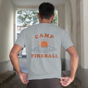 K&ouml;nnte beinhalten: Hellgraues T-Shirt mit dem orangefarbenen Schriftzug "CAMP FIREBALL" &uuml;ber einer Grafik von Sonne, Bergen und einer geometrischen Form. Das T-Shirt hat kurze &Auml;rmel und scheint aus einem weichen Material zu sein.