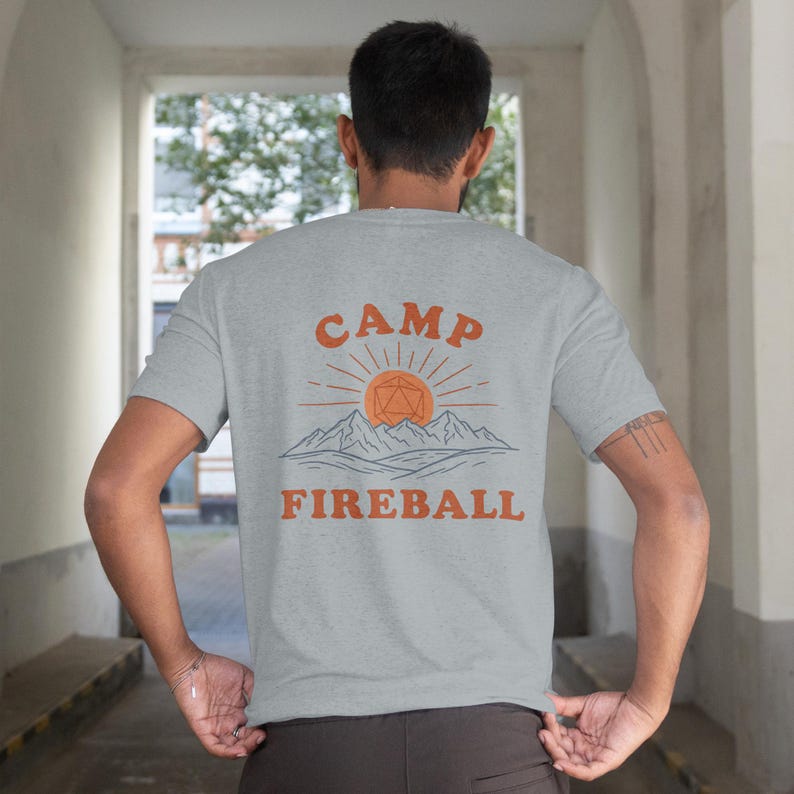 K&ouml;nnte beinhalten: Hellgraues T-Shirt mit dem orangefarbenen Schriftzug "CAMP FIREBALL" &uuml;ber einer Grafik von Sonne, Bergen und einer geometrischen Form. Das T-Shirt hat kurze &Auml;rmel und scheint aus einem weichen Material zu sein.