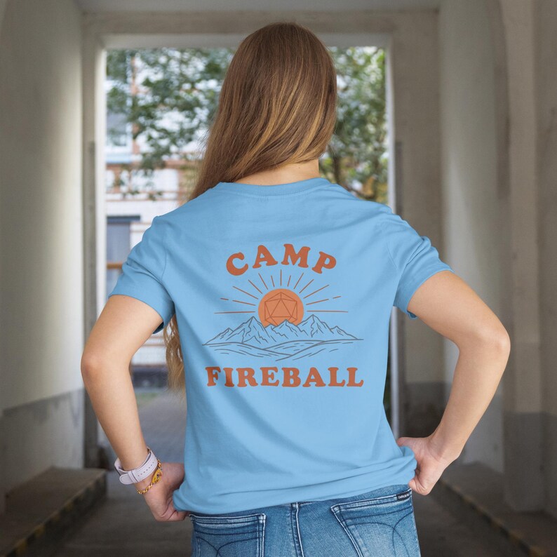 K&ouml;nnte beinhalten: Hellblaues T-Shirt mit dem orangefarbenen Schriftzug "CAMP FIREBALL" &uuml;ber einer Grafik von Sonne, Bergen und einer geometrischen Form. Das T-Shirt hat kurze &Auml;rmel und ist aus weichem Stoff.