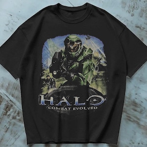 Könnte beinhalten: Ein schwarzes Kurzarm-T-Shirt mit einem Grafikdruck eines grün gepanzerten Soldaten aus dem Videospiel Halo Combat Evolved. Das Design enthält den Text Halo Combat Evolved in blauer und weißer Schrift unterhalb der Figur.