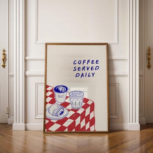 May include: A framed print featuring a coffee-themed illustration. The artwork depicts two coffee cups, a croissant, and the text "COFFEE SERVED DAILY" in blue. The background is a red and white checkered tablecloth.