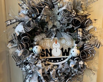 May include: A winter wreath with black, white, and silver ribbons, bows, and decorations. The wreath has a sign saying "Let it Snow" with a snowman and two small bells. The wreath is designed for the winter season.