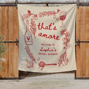 May include: A cream-colored fabric banner featuring red line-art illustrations of wine, herbs, and pasta. The text reads: that's amore, WELCOME TO Sophia's BRIDAL SHOWER. The design includes a wine bottle, a glass of wine, garlic, and sprigs of rosemary.