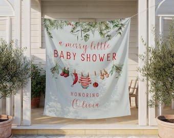 May include: A light blue fabric backdrop featuring a Christmas-themed illustration with green pine branches and red berries. The text reads a merry little BABY SHOWER HONORING Olivia. A clothesline displays red socks, a hat, a striped onesie, and mittens.