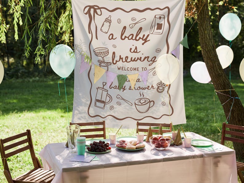 May include: A baby shower scene with a banner that reads "a baby is brewing welcome to a baby shower." The banner has coffee-themed illustrations. A table is set with food, drinks, and decorations, including balloons and a string of colorful flags.