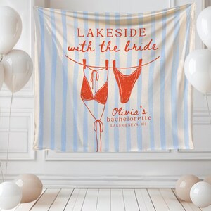 May include: A bachelorette party banner with the text "Lakeside with the bride" and a drawing of a red bikini. The banner is striped with blue and white. White balloons and decorative balls are in the foreground.