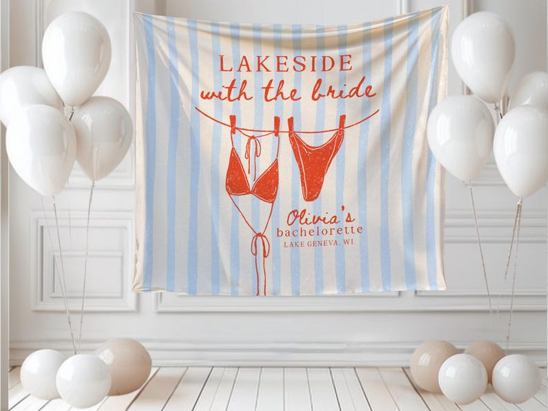 May include: A bachelorette party banner with the text "Lakeside with the bride" and a drawing of a red bikini. The banner is striped with blue and white. White balloons and decorative balls are in the foreground.