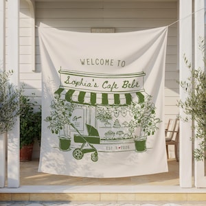 May include: A cream-colored banner with a green illustration of a cafe, a stroller, and the text "WELCOME TO Sophia's Cafe Bebe". The cafe has a striped awning and is surrounded by green plants. The banner is hanging outdoors.