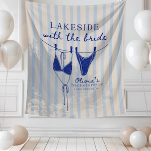 May include: A bachelorette party backdrop with blue and white striped design, featuring a drawing of a bikini. The text reads "Lakeside with the bride" and "Olivia's bachelorette Lake Geneva WI". White balloons and decorative spheres are in the foreground.