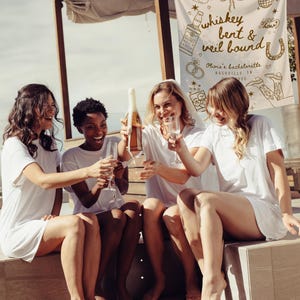May include: Four women in white t-shirts and shorts are celebrating outdoors. One is pouring champagne into glasses. A banner in the background reads "whiskey bent & veil bound" with Nashville, TN details, suggesting a bachelorette party.