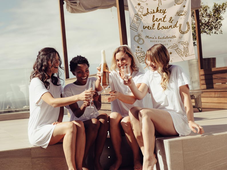 May include: Four women in white t-shirts and shorts are celebrating outdoors. One is pouring champagne into glasses. A banner in the background reads "whiskey bent & veil bound" with Nashville, TN details, suggesting a bachelorette party.