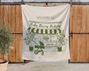 May include: A cream-colored fabric wall hanging featuring a green line-art illustration of a bakery storefront. The text reads WELCOME TO Baby Geary Bakery and EST 7 2026. A baby stroller sits in front of the shop, flanked by two potted citrus trees.