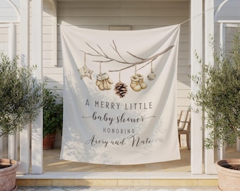 May include: A cream-coloured fabric banner featuring a watercolour illustration of a tree branch with hanging ornaments including a star, mittens, a pinecone, booties, and a ball. The text reads A MERRY LITTLE baby shower HONORING Avery and Nate.