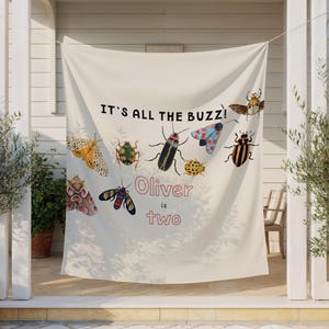 Puede incluir: Una pancarta color crema con el texto "IT'S ALL THE BUZZ!" y las palabras "Oliver is two". La pancarta presenta ilustraciones coloridas de varios insectos, incluyendo mariposas y escarabajos.