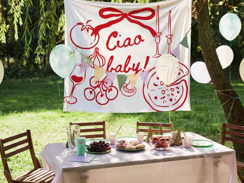 May include: A festive outdoor party scene with a white banner featuring red illustrations of Italian food and the phrase "Ciao baby!". A table is set with food, drinks, and plates. Balloons in various colors are scattered around the scene.