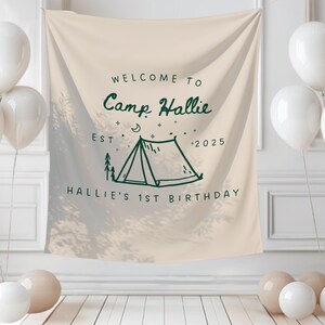 May include: A beige banner with the text "WELCOME TO Camp Hallie EST - 2025 HALLIE'S 1ST BIRTHDAY" in dark green. The banner features a tent illustration. White balloons are arranged around the banner, creating a festive atmosphere.