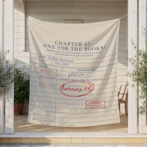 May include: A beige fabric banner with blue lines and text. The banner reads "CHAPTER 45: ONE FOR THE BOOKS!" with details like "Olivia Manning" and "est. 1981." A red circle surrounds "turning 45." The banner is displayed outdoors.