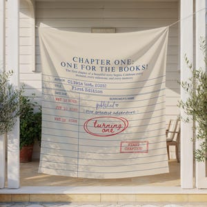 May include: A cream-coloured fabric banner with navy blue text reading "CHAPTER ONE: ONE FOR THE BOOKS!". The banner is designed to look like a library card, with printed lines and text, including "First Edition" and "turning one" in a red circle.