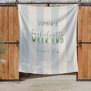 May include: A white and light blue striped banner with the text "SOPHIA'S bachelorette WEEK END miami. fl". The banner is hanging in front of a wooden door. A potted plant is on the left side of the image.