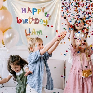 May include: A white fabric backdrop featuring the colorful text HAPPY 1st birthDAY LIAM, displayed alongside a cluster of peach, gold, and white balloons, with confetti falling over children in a room setting during a birthday celebration.