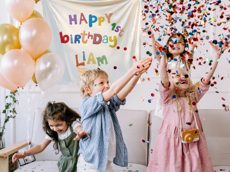 May include: A white fabric backdrop featuring the colorful text HAPPY 1st birthDAY LIAM, displayed alongside a cluster of peach, gold, and white balloons, with confetti falling over children in a room setting during a birthday celebration.