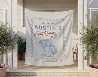 Może przedstawiać: Kremowe tło z tkaniny z niebieską obwódką w stylu liny. Napis AUSTIN'S First Rodeo oraz SADDLE UP! wykonano w kolorze niebieskim i czerwonym. Niebieski rysunek liniowy przedstawia osobę ujeżdżającą wierzgającego konia wewnątrz ramki.