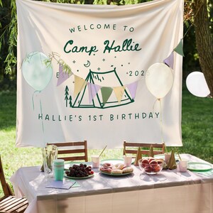 May include: A festive outdoor birthday party scene. A banner reads "Welcome to Camp Hallie" with a tent illustration and "Hallie's 1st Birthday". Balloons and a table set with food and drinks complete the celebration.