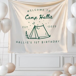 May include: A cream-colored banner with the text "WELCOME TO Camp Hallie" and "HALLIE'S 1ST BIRTHDAY" in green. A green tent graphic is centered on the banner. White and tan balloons are arranged around the banner.