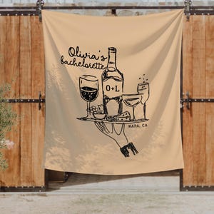 K&ouml;nnte beinhalten: Beige Stoffbanner mit einer schwarzen Illustration einer Weinflasche, Gl&auml;sern und Essen auf einem Tablett, gehalten von einer Hand. Der Text lautet "Olivia's bachelorette" und "NAPA, CA".