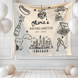 May include: A cream-colored banner with black line drawings of Chicago landmarks, food, and celebratory items. The banner reads "Olivia's Bachelorette" and "Chicago". White balloons and a light-colored floor complete the scene.