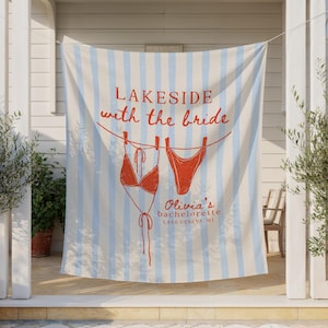 May include: A beach towel with blue and white vertical stripes. The towel has a red illustration of a bikini top and bottom hanging on a clothesline. Text reads "Lakeside with the Bride" and "Olivia's bachelorette Lake Geneva WI."
