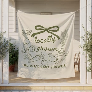 Może przedstawiać: Kremowy baner z zieloną kokardą i ilustracjami warzyw. Tekst brzmi "locally grown" i "Olivia's Baby Shower". Baner wisi między dwoma doniczkowymi drzewami oliwnymi.