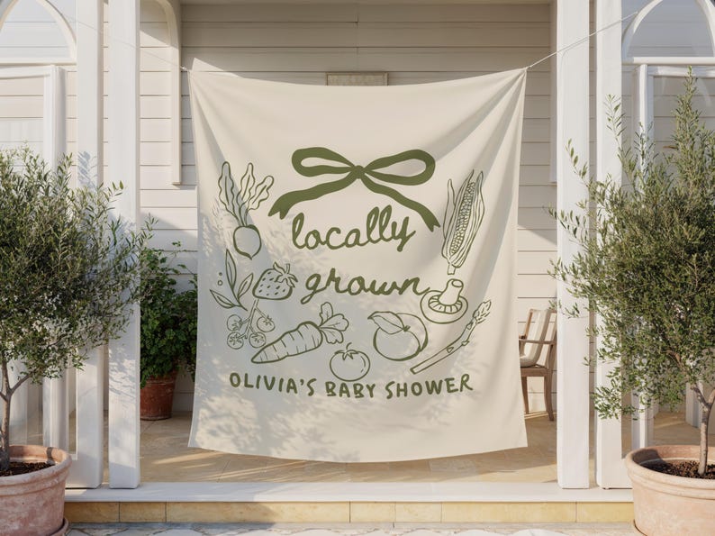 Może przedstawiać: Kremowy baner z zieloną kokardą i ilustracjami warzyw. Tekst brzmi "locally grown" i "Olivia's Baby Shower". Baner wisi między dwoma doniczkowymi drzewami oliwnymi.