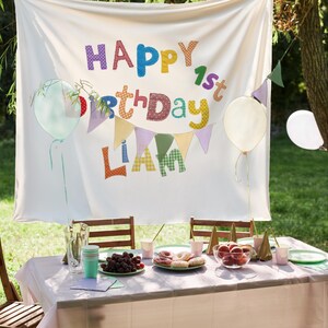 May include: A white fabric backdrop featuring the colorful text HAPPY 1st BIRTHDAY LIAM with a bunting banner. Below, a table is set with a light pink tablecloth, plates of grapes and donuts, a bowl of peaches, cups, and a pitcher.