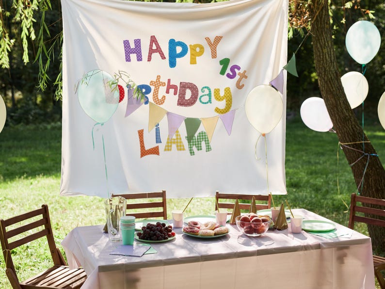 May include: A white fabric backdrop featuring the colorful text HAPPY 1st BIRTHDAY LIAM with a bunting banner. Below, a table is set with a light pink tablecloth, plates of grapes and donuts, a bowl of peaches, cups, and a pitcher.