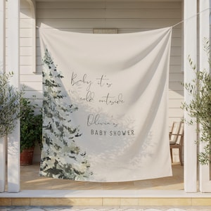 May include: A cream-colored baby shower banner with a winter theme. The banner features a watercolor-style evergreen tree and the text "Baby it's cold outside" and "Olivia's BABY SHOWER" in elegant script.