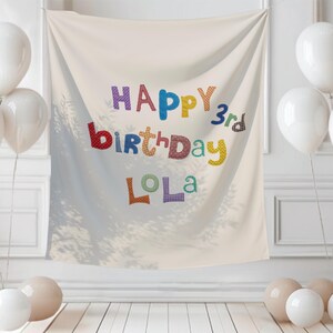 May include: A cream-colored fabric backdrop featuring the text HAPPY 3rd birthday LOLA in multi-colored, patterned letters. The banner is surrounded by clusters of white and beige balloons on both sides, creating a festive display.