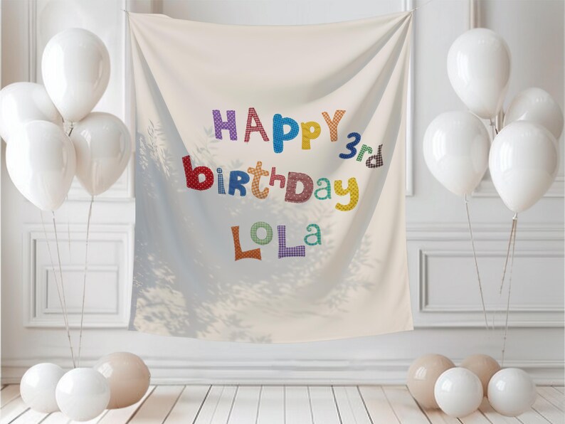 May include: A cream-colored fabric backdrop featuring the text HAPPY 3rd birthday LOLA in multi-colored, patterned letters. The banner is surrounded by clusters of white and beige balloons on both sides, creating a festive display.