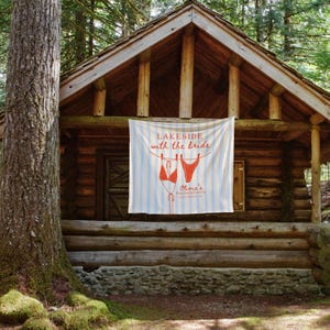 May include: A rustic log cabin in a forest setting. A banner hangs from the porch with the text "Lakeside with the Bride" and a bikini graphic. The banner has blue and white stripes. The cabin is surrounded by trees.