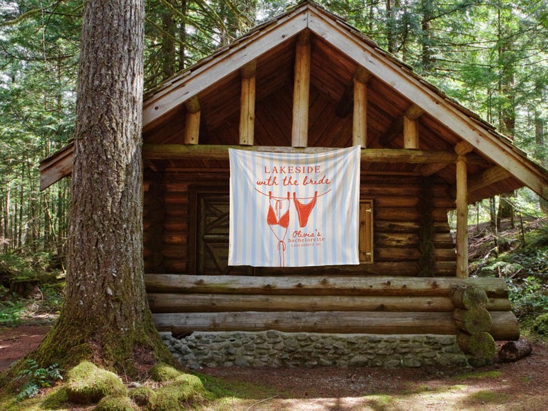 May include: A rustic log cabin in a forest setting. A banner hangs from the porch with the text "Lakeside with the Bride" and a bikini graphic. The banner has blue and white stripes. The cabin is surrounded by trees.