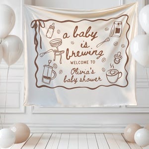 May include: A white banner with brown illustrations and text that reads "a baby is brewing" and "Welcome to Olivia's baby shower." The banner is surrounded by white balloons and beige orbs.