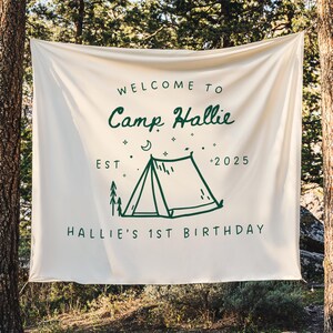 May include: A cream-colored banner with the text "WELCOME TO Camp Hallie" in a whimsical green font. Below, a green illustration of a tent, trees, and stars, with the text "EST 2025" and "HALLIE'S 1ST BIRTHDAY". The banner is hanging between two trees.