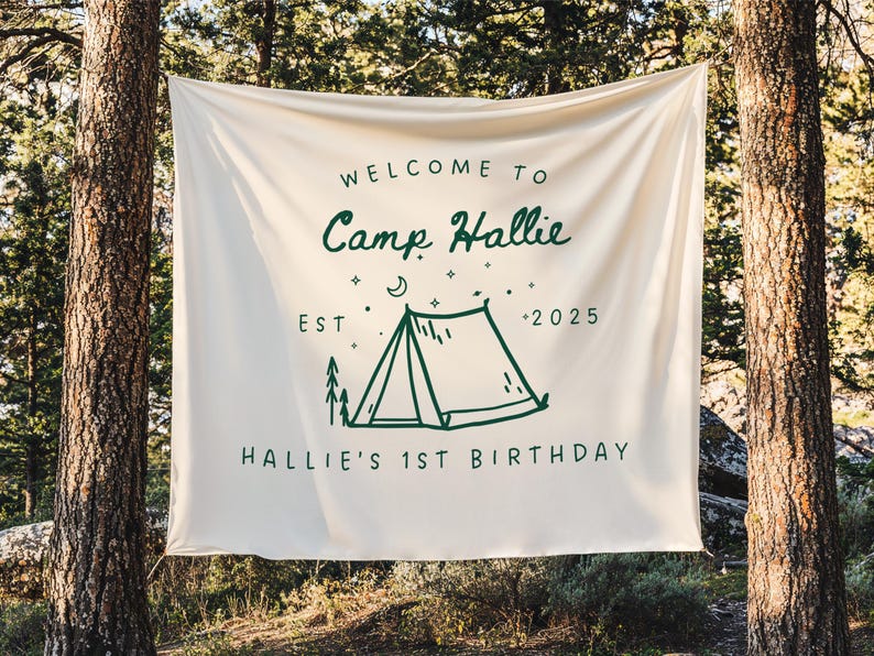 May include: A cream-colored banner with the text "WELCOME TO Camp Hallie" in a whimsical green font. Below, a green illustration of a tent, trees, and stars, with the text "EST 2025" and "HALLIE'S 1ST BIRTHDAY". The banner is hanging between two trees.